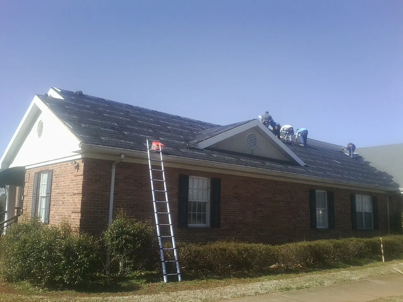 Crew tearing shingles off a brick ranch roof with a ladder set — Emergency Roof Repair work by our crew in Ambrose