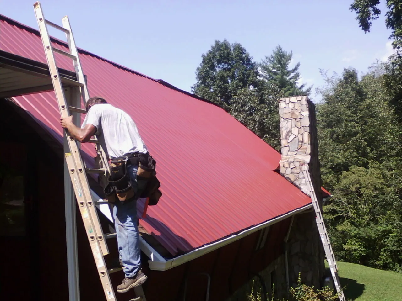 Roofer on a ladder beside a red metal roof and stone chimney — Soffit & Fascia Repair by Roofing Brothers in Ambrose